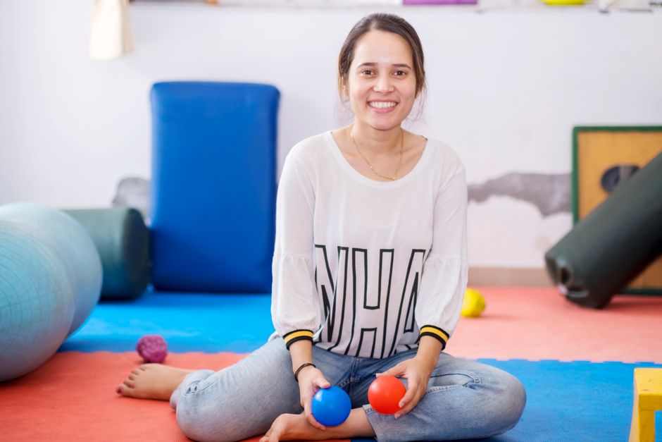 Smiling woman sitting cross-legged on colorful exercise mats, holding a blue and a red ball.