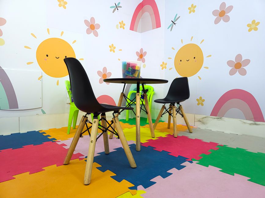 Child-friendly play area with a small round table and two black chairs on a colorful foam mat; walls feature sun, rainbow, and flower decor.