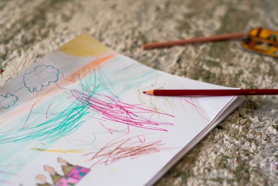 Child's colorful crayon drawing on notebook laid on a rough outdoor surface; pencils and a measuring tape nearby.