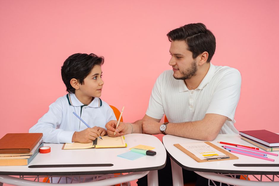 Adult man and boy sit at a desk, studying together with notebooks and pencils on a pink background.