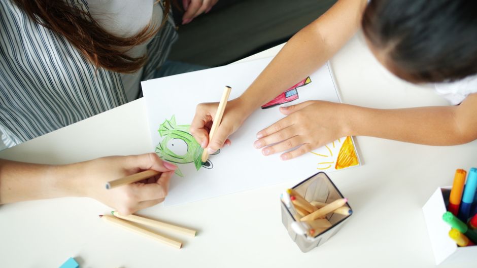 Children drawing colorful pictures at a white table with pencils and markers nearby, creating a green creature and a yellow sun on a sheet of paper.