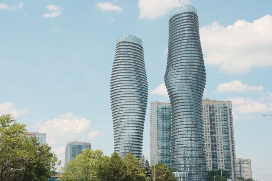 Twin twisting glass skyscrapers rise above trees and city street on a sunny day.