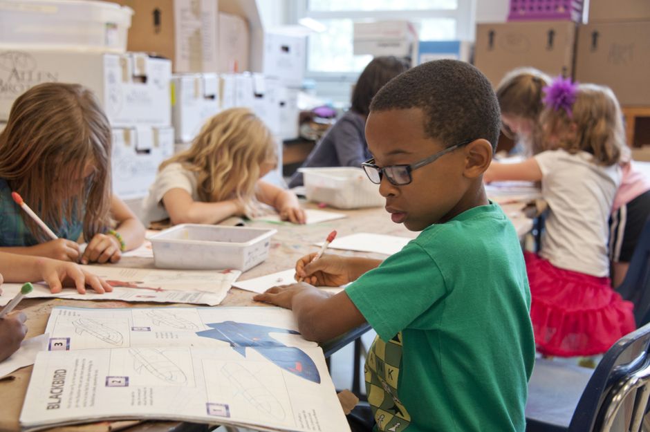 Children at a classroom table drawing and coloring with pencils, focused on worksheets and supplies around them