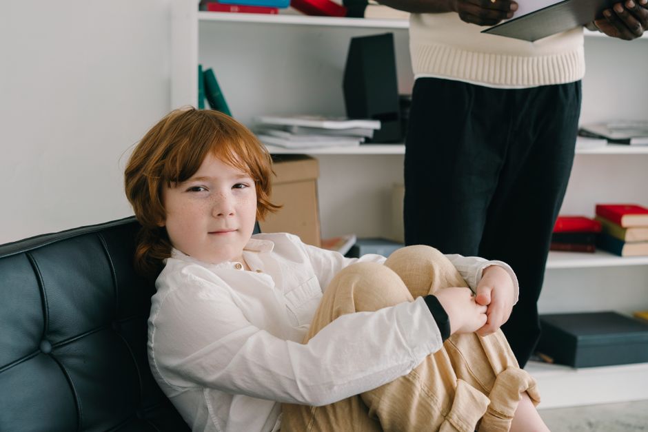 A young girl with red hair sits on a black leather sofa, knees drawn up, looking at the camera, while an adult stands nearby holding a clipboard in a room with shelves and books.