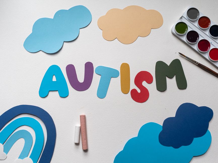 Colorful cutout letters spell 'AUTISM' on a white surface with clouds and a paint set nearby.