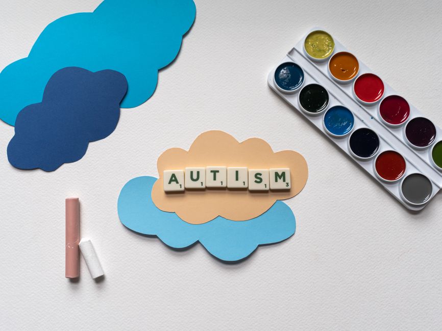 Art supplies and cloud shapes with Scrabble letters spelling AUTISM, symbolizing autism awareness and creativity.