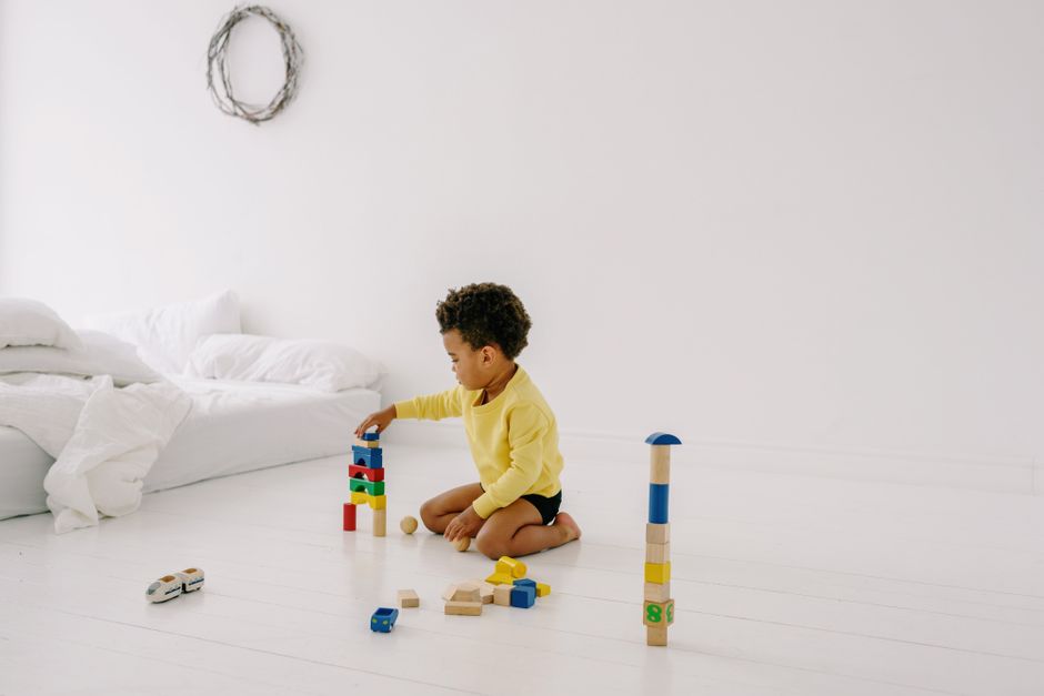 A young child wearing a yellow shirt sits on a white floor, stacking colorful wooden blocks beside a bed in a bright room.