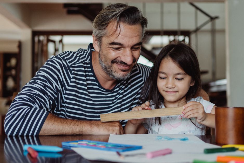 Man and young girl at a table drawing with a wooden ruler, both smiling as they work on a colorful art project.